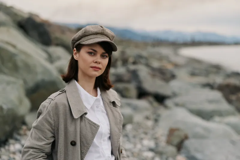 Young woman wearing a textured coat and matching cap stands on rocky beach with blurred background. The natural setting and casual outfit create a calm and contemplative outdoor portrait.