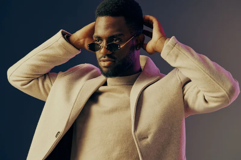 Stylish young man adjusting his sunglasses, wearing a trendy coat, posing against a vibrant background, showcasing contemporary fashion and confidence