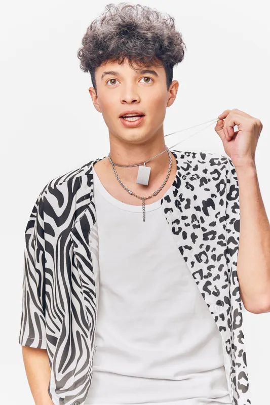 Young man with curly hair wearing a black and white patterned shirt and white tank top, holding a necklace against a clean white background.
