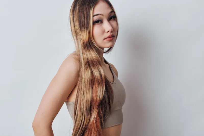 Young woman with long straight hair wearing a beige tank top posing against a light background looking at camera with a neutral expression in studio portrait.