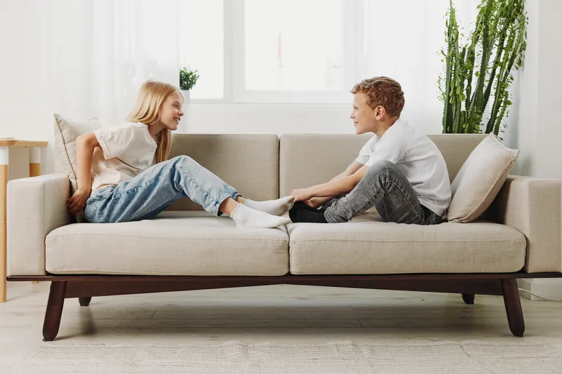 Happy boy and girl sitting on beige sofa playing footsies in bright living room with modern decor and large window. Concept of childhood fun, friendship, and cozy home atmosphere.