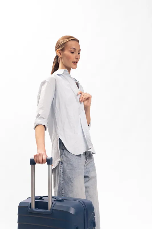 Young woman in casual clothes standing with a navy suitcase on white background. Travel concept with female traveler holding luggage in studio. Minimalist style and relaxed mood.