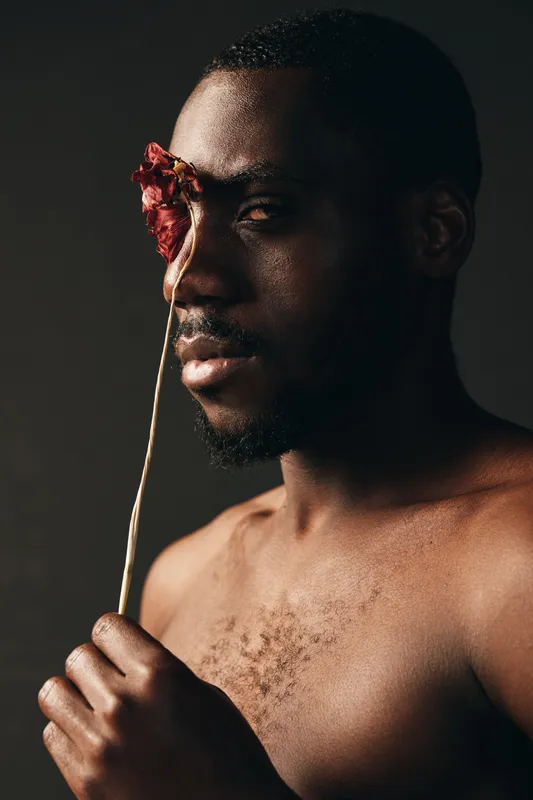 Creative conceptual portrait of African man with dark skin holding dried flower in front of face, artistic visual style with moody lighting and bare chest for emotional expression.