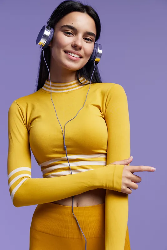 sport fitness woman wearing yellow activewear and headphones, smiling confidently against purple background in indoor studio lighting