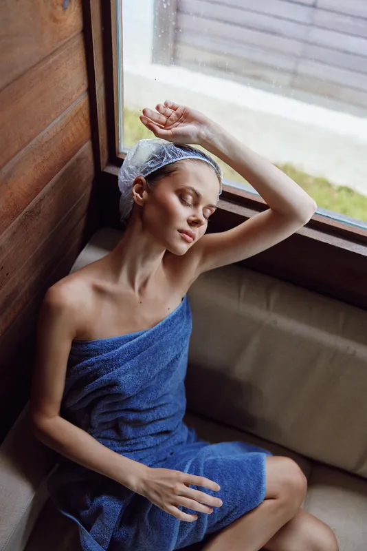 woman relaxing in sauna wrapped in blue towel with shower cap on head, natural light through window, serene expression, wooden interior background