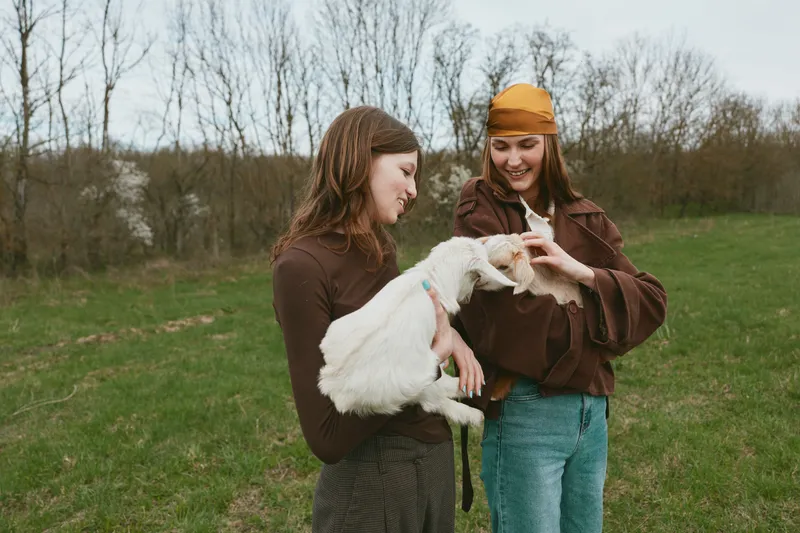Two friends outdoors with a playful goat kid, smiling and handling the animal, casual jackets and jeans, relaxed countryside lifestyle moment on a green field.