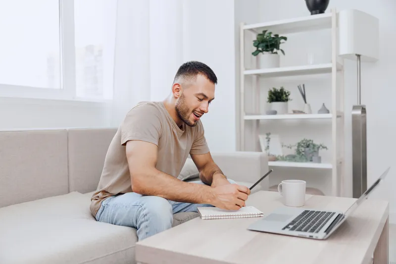 Focused man writing in a notebook while sitting on a cozy couch, surrounded by natural light. Ideal for themes of productivity and home office inspiration.
