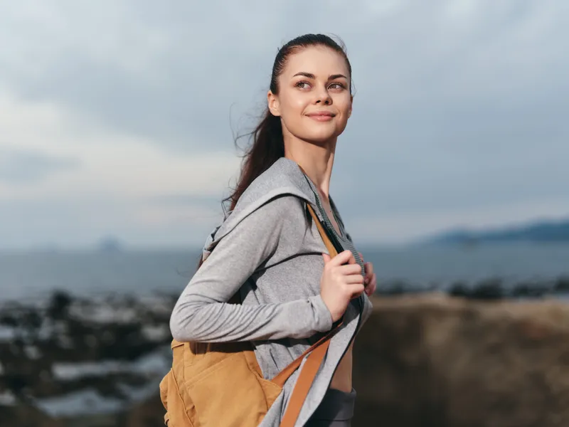 Young woman with a backpack smiling at the beach, dressed in casual sportswear, showcasing confidence and adventure against a cloudy sky