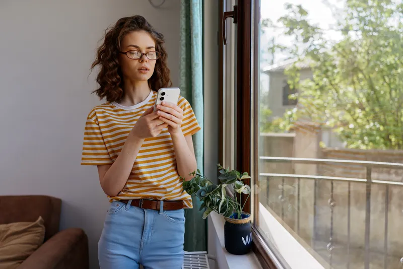 Soft daylight pours through a sunlit window as a young woman with curly brunette hair checks her phone, standing by a potted plant in a tidy, modern living space.