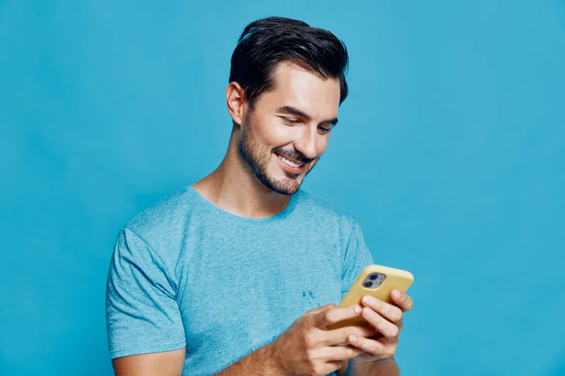 Smiling man using mobile phone, dressed in casual blue t-shirt, enjoys communication while isolated on bright azure background, expressing happiness and connection.