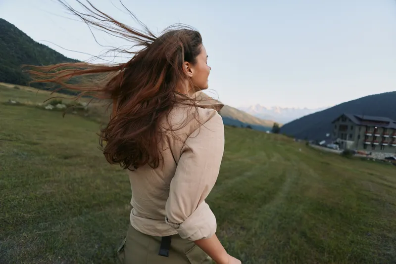 Young brunette woman with long hair enjoys a windy day outdoors in casual beige jacket while looking at distant mountains in a green open field.