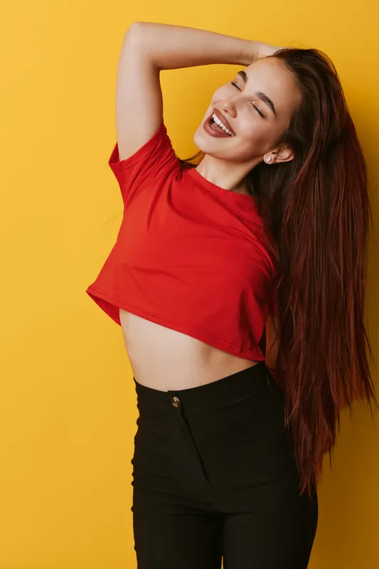 Young woman with long hair wearing red crop top and black pants smiling with eyes closed posing on yellow background. Happy female model expressing joy and confidence in casual outfit.