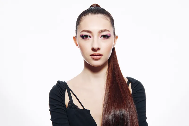 Young woman with glossy long brown hair and creative pink eye makeup, wearing black stylish top, posing against a white seamless background. Bright beauty fashion portrait.