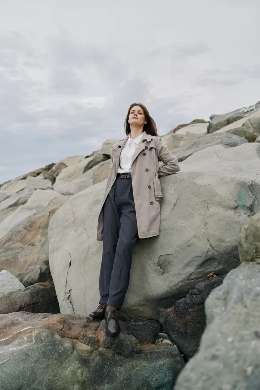 Young woman in a stylish coat and black pants leans on large rocks at the coast. Overcast sky and natural stone background create a calm and thoughtful mood.