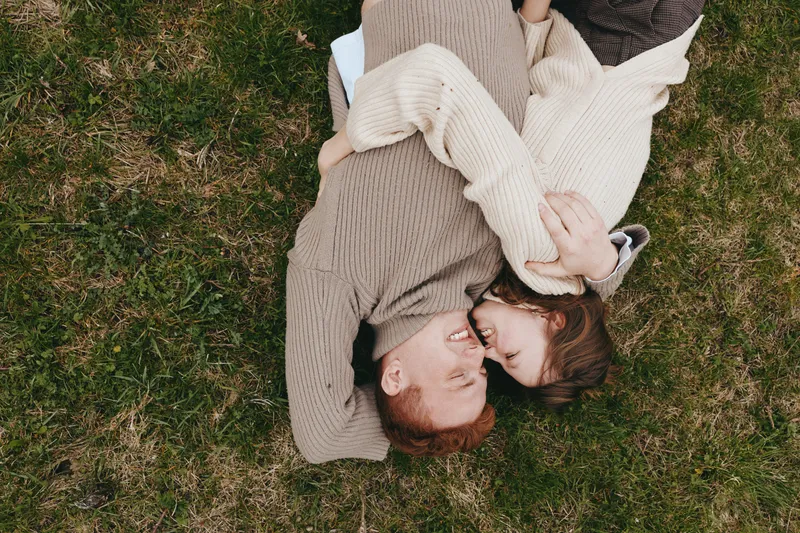A couple lies on a grassy field, cuddling and smiling, wearing knit sweaters in soft daylight. An intimate, relaxed moment outdoors expressing affection, connection, and everyday lifestyle.