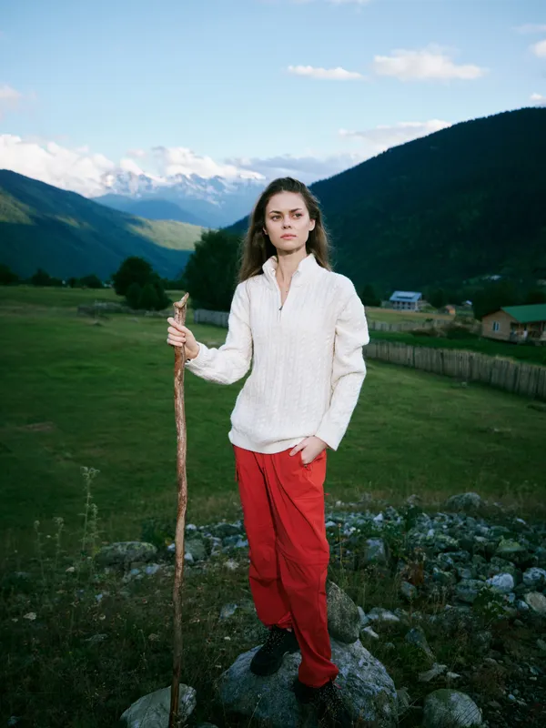 Hiking outdoor scene with a woman standing on a rocky field holding a staff, mountains and valley in the background, wearing a white sweater and red pants, rustic landscape