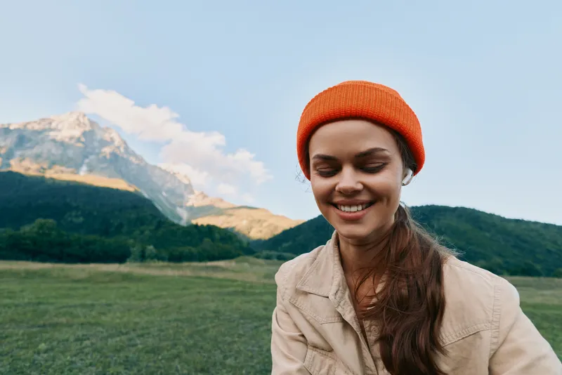 A smiling woman in a beige jacket and orange beanie enjoys a sunny outdoor moment in a green meadow with distant mountains, capturing a relaxed, casual lifestyle.