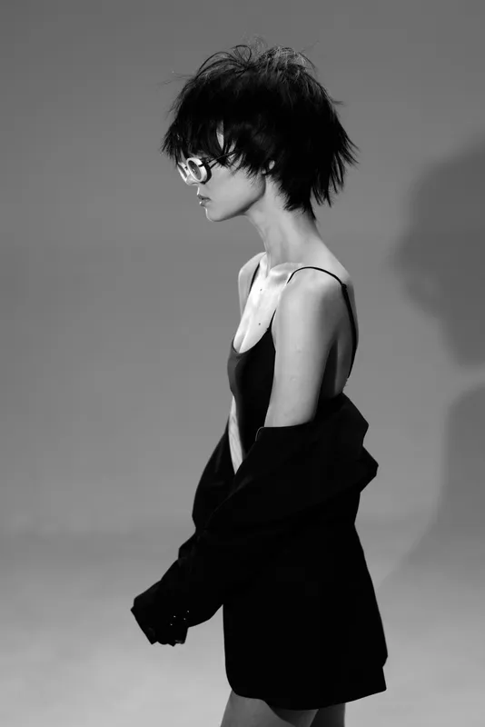 Black and white portrait of a stylish woman with short hair wearing glasses and a black outfit, showcasing modern fashion and minimalistic elegance.