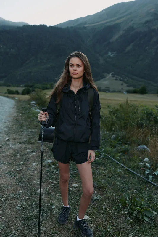 A young woman stands on a rocky trail in a mountain landscape, holding a trekking pole. She wears a dark jacket and shorts, focused and ready for adventure.