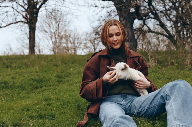 Calm woman outdoors, sitting on green grass in a park, holding a playful goat kid, gentle smile, cozy jacket and jeans, natural light, peaceful setting, candid lifestyle moment