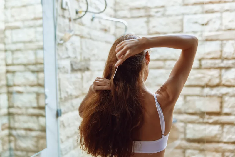 Woman with long hair combing in the bathroom, natural light, relaxed morning routine, casual white top, minimalistic interior design.