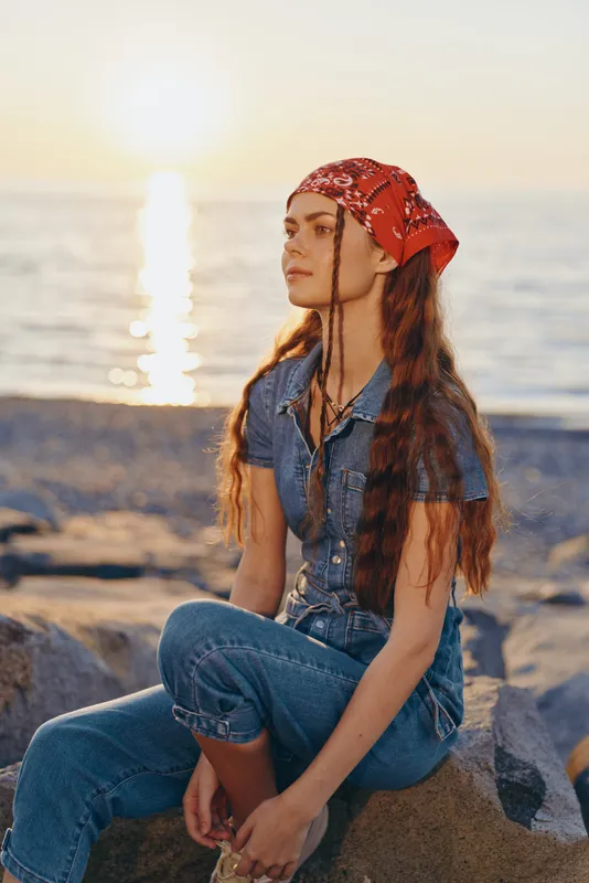 Lifestyle woman in modern boho-western denim street style sits by the water during golden hour with film color warmth and relaxed, thoughtful expression.