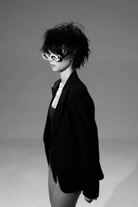 Black and white portrait of woman wearing sunglasses and blazer with tousled hair in studio lighting.