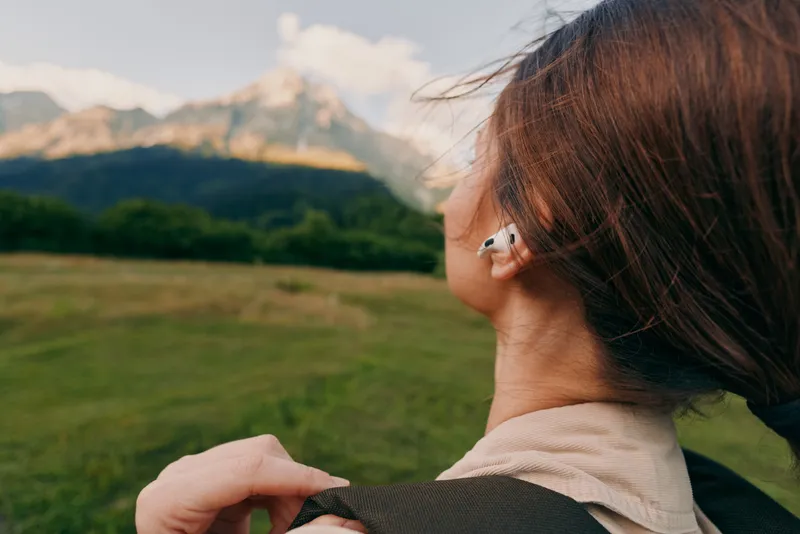 A woman seen from behind with a backpack in an open field, mountains in the distance and wind in her hair, capturing outdoor lifestyle.
