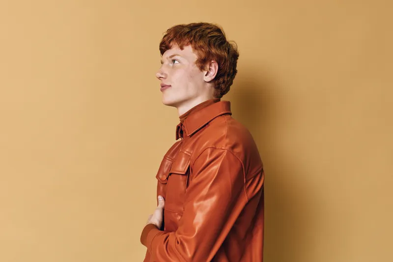 Young man in leather jacket standing in profile against orange background with arms crossed looking thoughtful confident casual fashion style portrait.