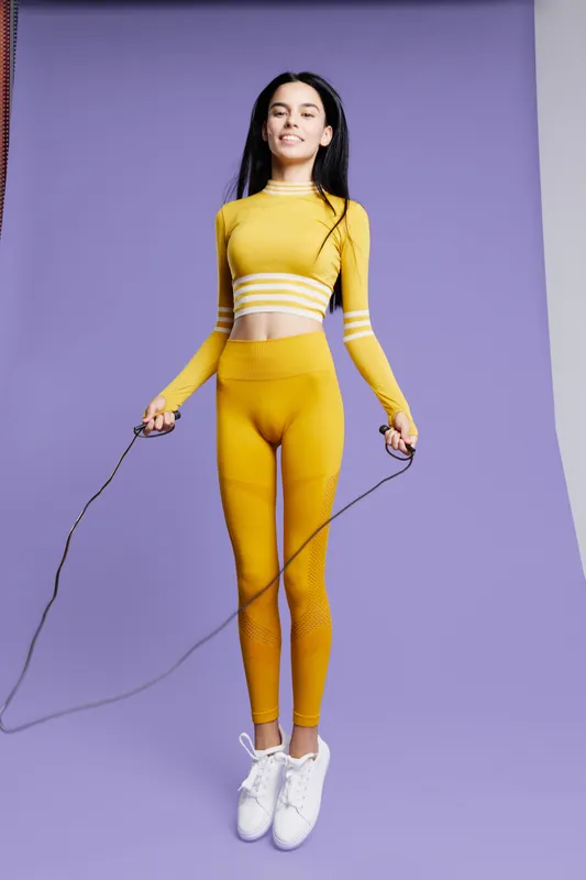 young woman sport fitness jumping rope wearing bright yellow activewear with white sneakers on purple background, energetic and focused workout moment