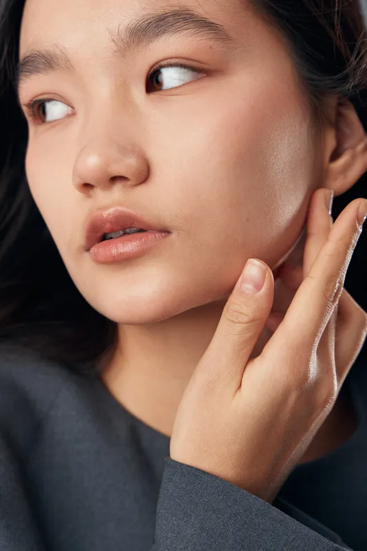 Portrait of young woman touching face with hand wearing dark top, natural skin, thoughtful expression and looking aside against dark background in studio lighting.
