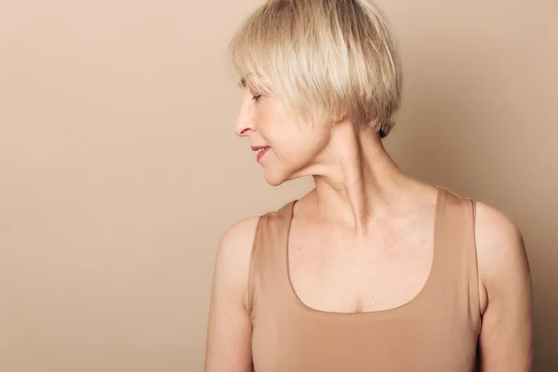 Profile of mature woman with short blonde hair wearing sleeveless top looking sideways on beige background. Concept for aging, beauty, lifestyle, confidence, and natural appearance.