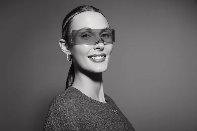 A smiling young white woman with light skin wears large futuristic sunglasses and a textured sweater. Studio portrait on grey background with modern fashion concept.