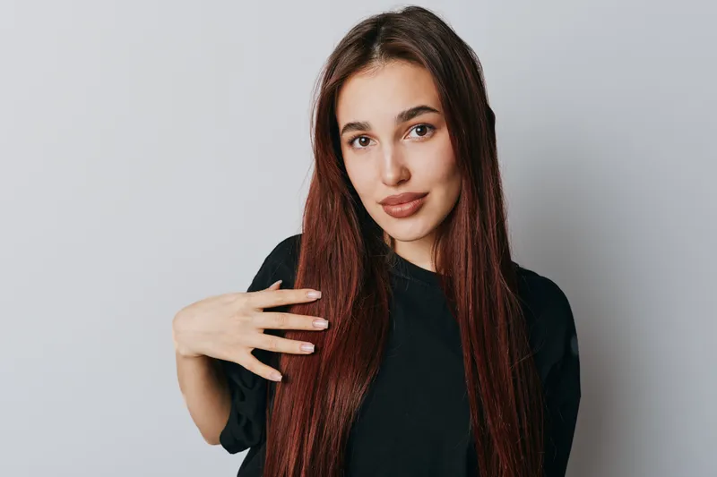 Young woman with long straight hair and natural makeup wearing black shirt posing with hand near face against plain light background. Beauty, fashion and casual style portrait.