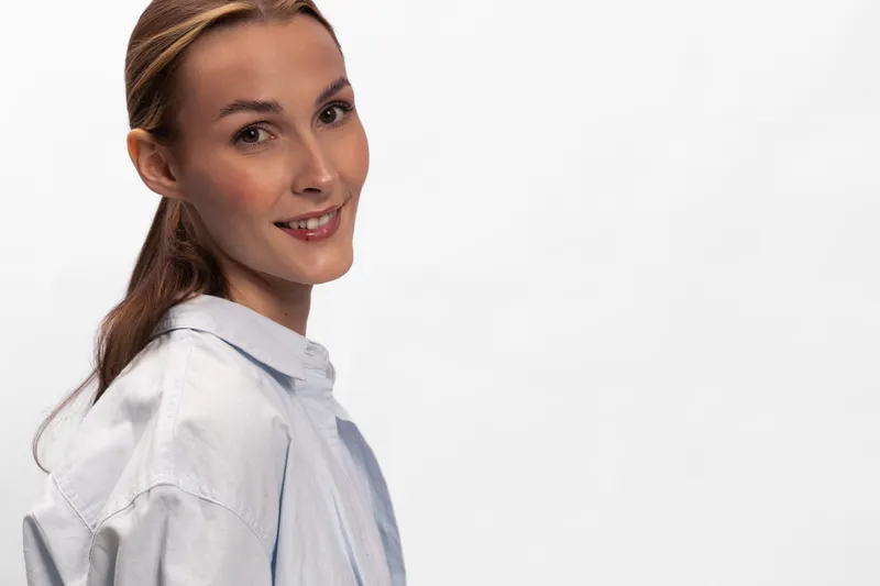 Smiling young woman with long brown hair wearing a light shirt looks at camera, isolated on white background. Concept for beauty, portrait, confidence, and casual style.