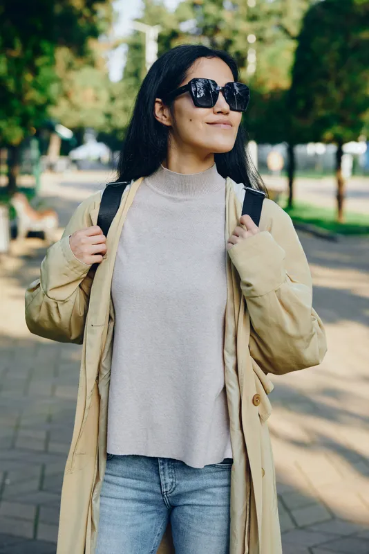 Sunglasses wearing woman in a beige coat and knit sweater strolling through a sunny park for casual outdoor fashion and confident relaxed vibe