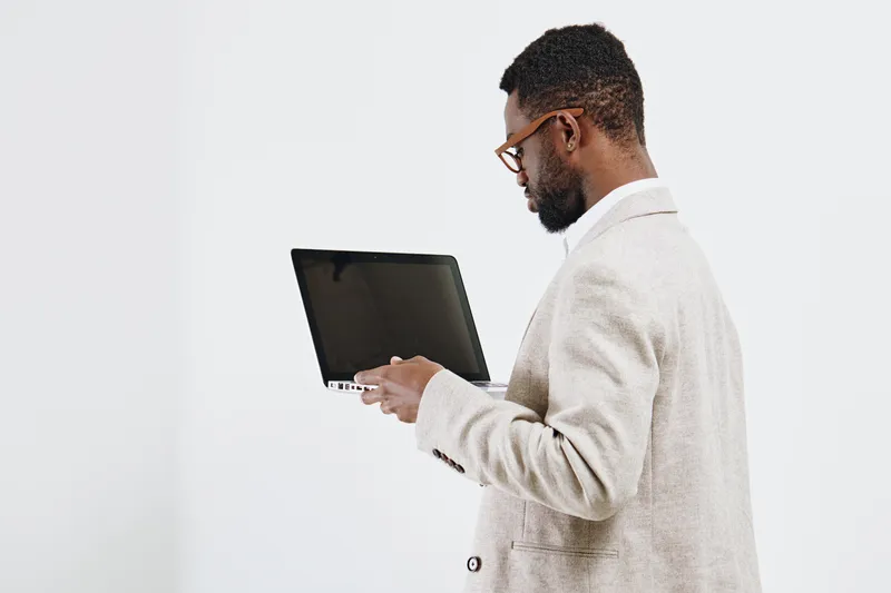 Professional young man with beard and glasses wearing beige suit holding a laptop computer while standing against a plain white background in a studio shot, business and technology concept.