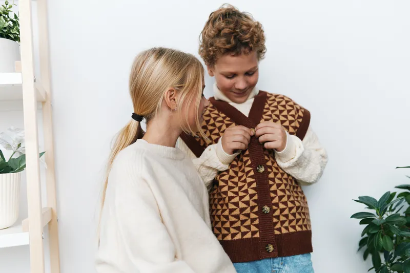 Authentic moment of youth at play in a cozy setting, capturing a school lifestyle scene as friends share a knit cardigan and engage in candid, gentle interaction between light walls and soft textures.