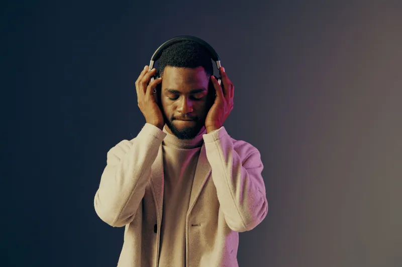 Young Black man enjoying music with headphones, wearing a stylish light sweater, against a gradient background showcasing calming colors, embodying tranquility and relaxation