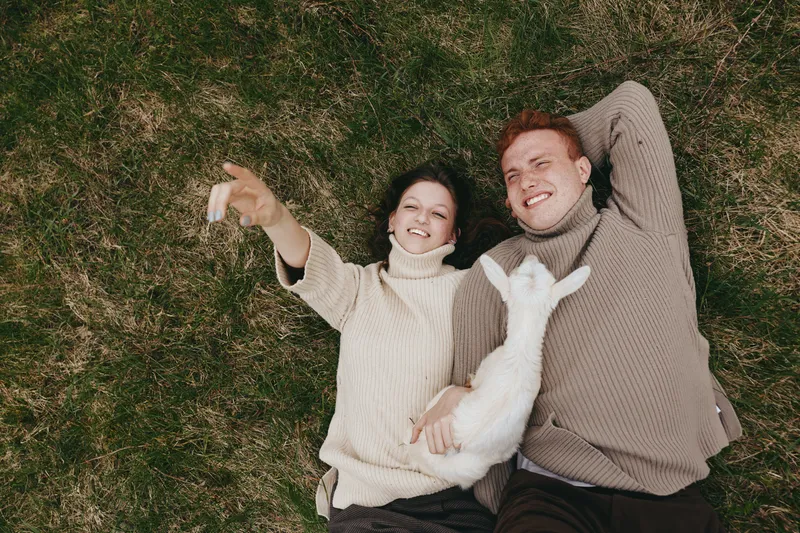 Young couple lying on grass with a white baby goat enjoying a relaxed moment together in cozy sweaters during a calm outdoor lifestyle scene.