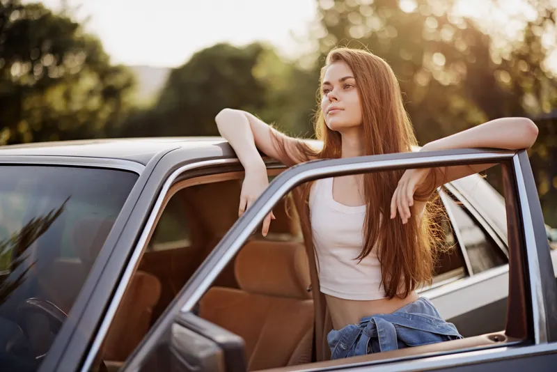 Young woman with long hair in a car, enjoying sunset, casual outfit, nature background, reflecting freedom and relaxation in summer vibes.