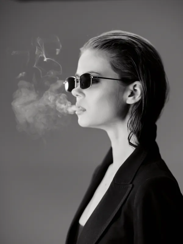 Black and white portrait of a stylish woman wearing sunglasses exhaling smoke in profile with sleek hair and dark blazer.
