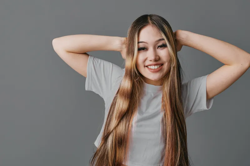 Young woman with long straight hair wearing gray t shirt smiling and holding hands behind head posing in studio with plain gray background for beauty and happiness concept.