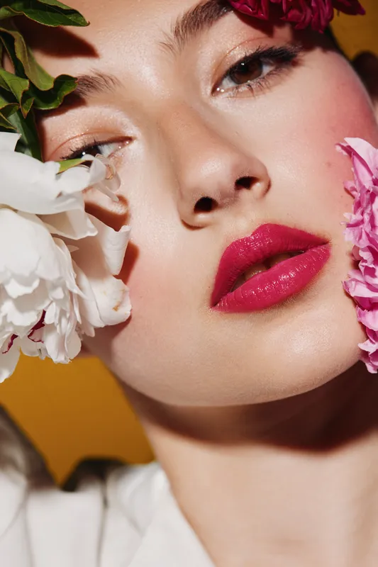 Close-up of a woman with glossy pink lips and dewy skin surrounded by white and pink flowers. Soft lighting accentuates creative beauty and fashion concept with a bold yellow background.