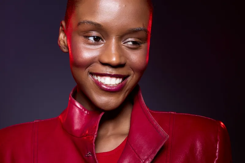 Close-up of a smiling dark skinned African woman wearing a red leather jacket. Studio portrait with soft lighting on a dark background. Concept of confidence, fashion, and beauty.