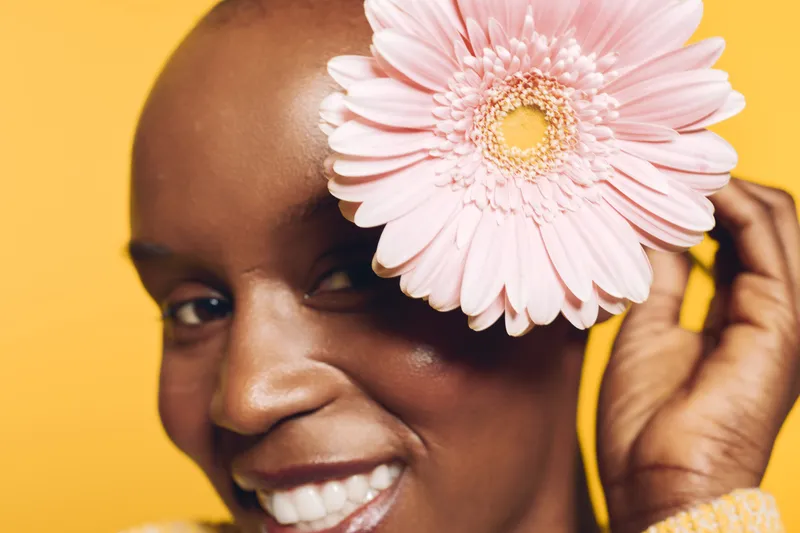 Close-up of happy dark skinned African woman smiling with a pink flower near her face on yellow background. Concept of beauty, natural skin, floral, and positive mood.