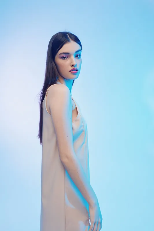 Young woman with long dark hair wearing a beige dress poses against a blue gradient background. Soft lighting highlights her beauty and fashion style in a studio setting.