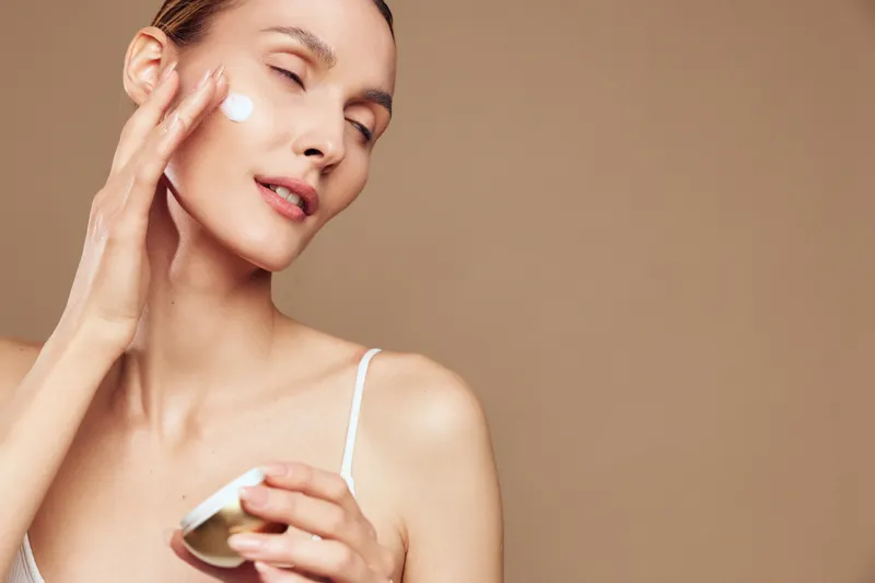 Young woman with clear skin applying white face cream on cheek with eyes closed on beige background. Concept of skincare, beauty, wellness, and self-care with soft natural lighting.