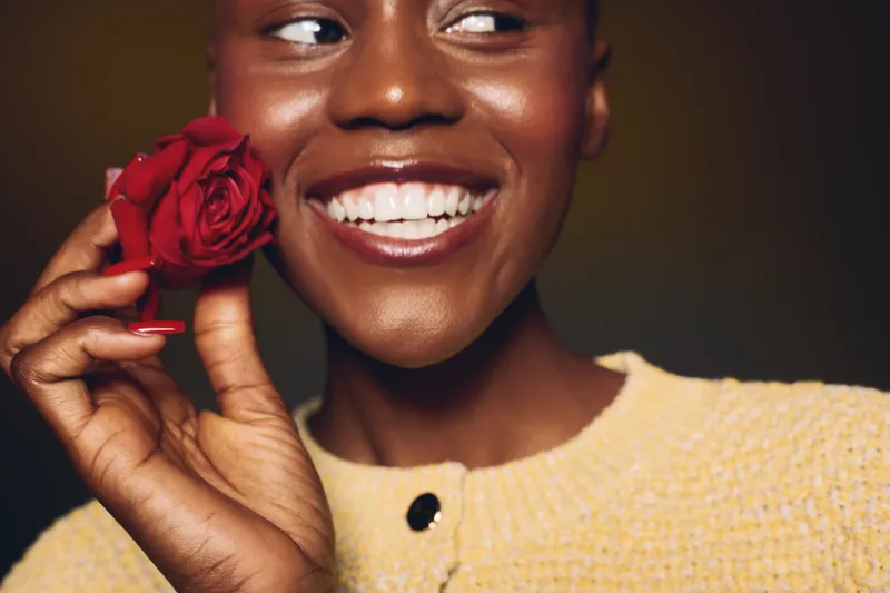Happy African woman with dark skin holds red rose near face, smiling and looking away. Close-up portrait on dark background. Concept of beauty, joy, and natural skincare.