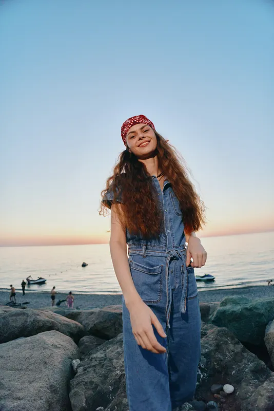 lifestyle woman in modern boho-western denim outfit smiling by the seaside at sunset with film color tones and relaxed street style vibes